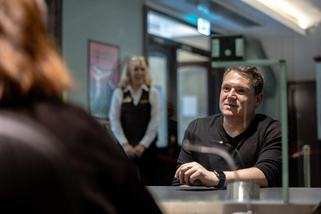 A white man using a wheelchair talks to a staff member at a theatre box office