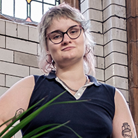 A white person with dyed blonde hair and glasses wears a blue top and jeans. Beth is wearing silver earrings, a necklace, and has tattoos on their arms.