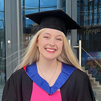 A white person with long blonde hair. Ceri-Anne is wearing a university academic cap with robes over a pink top.