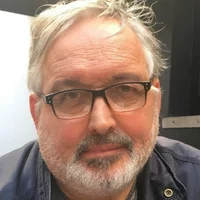 A white person with short silver hair and a short silver and grey beard. Graham has black and brown glasses and is wearing a blue jacket.