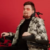 A white person with red hair and a red beard in a wheelchair against a bright red background. Jamie is wearing bright red eyeshadow and is wearing a black jumper and trousers with a white and grey blazer with black flowers.