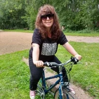 A white person with curly brown hair wearing sunglasses. Jules is sat on a blue bike and is wearing a black t-shirt, black trousers, and red Converse.