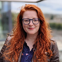 A white person with long, ginger hair and black glasses. Julie is wearing a blue floral shirt under a brown jacket.