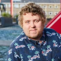 A white person with curly dark blond hair and a partial beard. Paul is wearing a blue shirt with palm tree patterns.