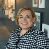 A white person with short blonde hair. Sam is wearing a black and white chequered shirt and a silver necklace.