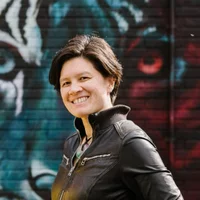 A Chinese-Caucasian biracial person with short dark hair. Shannon is wearing a black jacket with silver zips, and a clear and green necklace.