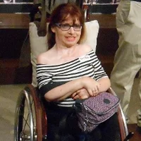 A white person with straight red hair in a wheelchair. Sophie has black glasses and is wearing a black and white striped off the shoulder top with cropped black trousers and a patterned pink bag.