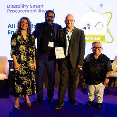 Abid Hussain a Roger Dennis yn derbyn Gwobr Gaffael Disability Smart ar ran All In a Chyngor Celfyddydau Lloegr ar lwyfan sydd wedi ei oleuo’n borffor yn Seremoni Wobrwyo Disablity Smart 2025. Mae Diane Lightfoot, Prif Swyddog Gweithredol y Fforwm Busnes Anabledd, a Simon Minty, llysgennad y Fforwm Busnes Anabledd, yn sefyll bob ochr iddynt ar y llwyfan.