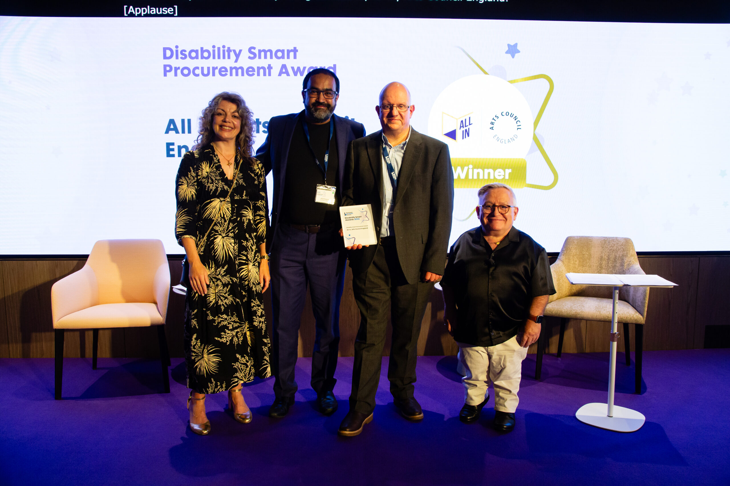 Abid Hussain a Roger Dennis yn derbyn Gwobr Gaffael Disability Smart ar ran All In a Chyngor Celfyddydau Lloegr ar lwyfan sydd wedi ei oleuo’n borffor yn Seremoni Wobrwyo Disablity Smart 2025. Mae Diane Lightfoot, Prif Swyddog Gweithredol y Fforwm Busnes Anabledd, a Simon Minty, llysgennad y Fforwm Busnes Anabledd, yn sefyll bob ochr iddynt ar y llwyfan.