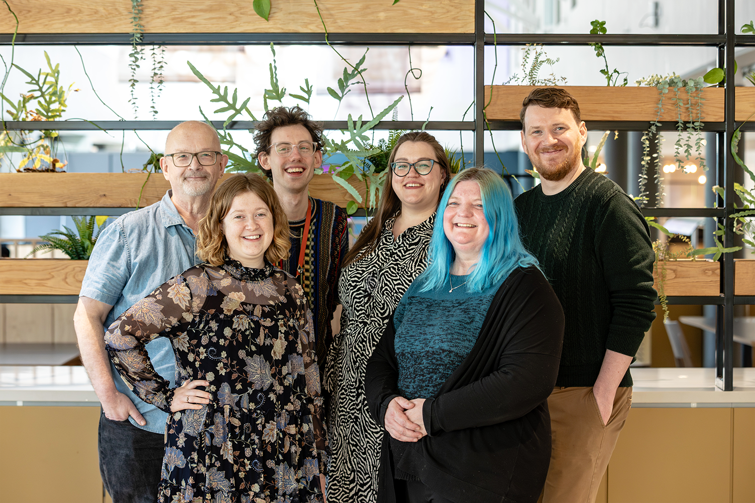 The All In team smile as they pose for a photo in a bright environment with plants and modern decor.