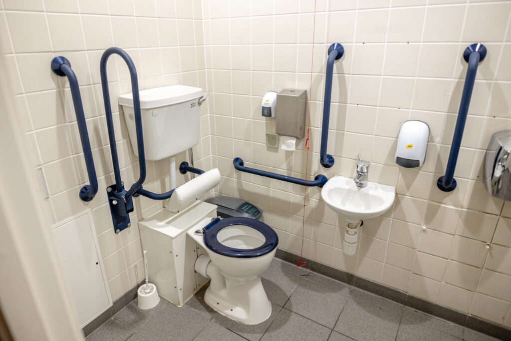 An accessible toilet showing the placement of the toilet, grab rails in contrasting colours, knee clearance, sink position, and emergency pull cord.