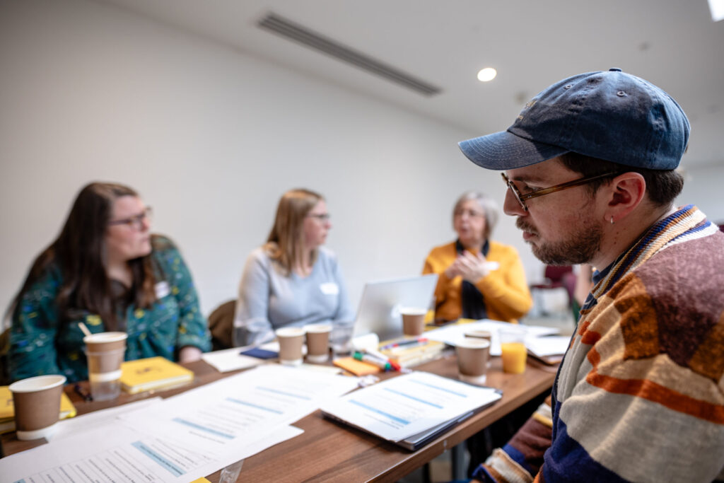 A group of people sit around a table having a discussion. In the foreground, someone wearing a colourful jumper and a baseball cap looks thoughtful.