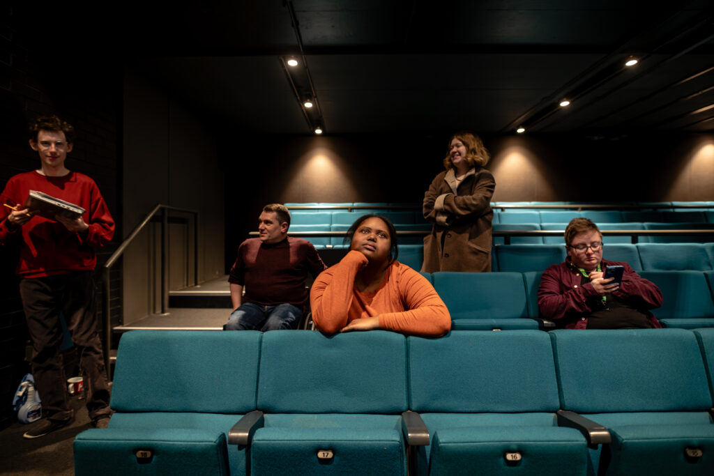 People seated and standing in a small auditorium with teal-colored seats and dark walls.