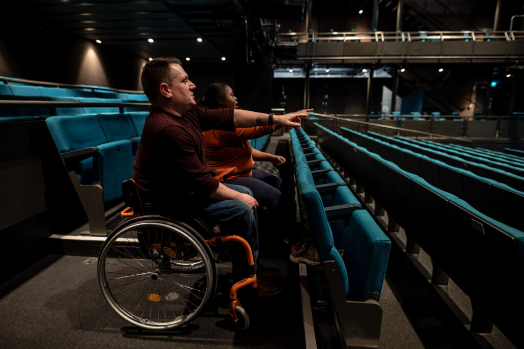 A group of audience members seated in a theatre auditorium admiring their view. One person is using a wheelchair space.