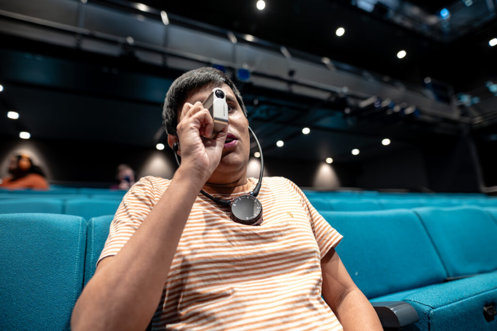 Person seated in a theatre with teal seats, wearing a striped shirt and an audio description device around their neck. They are holding a magnifying device.