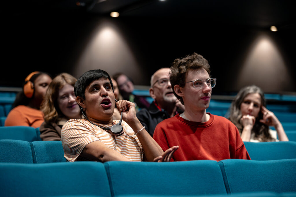 Several people are seated in a theatre with teal-coloured seats, watching something on stage or screen. One person is wearing an audio description receiver on a neck loop. In the background, someone is wearing bright orange ear defenders.