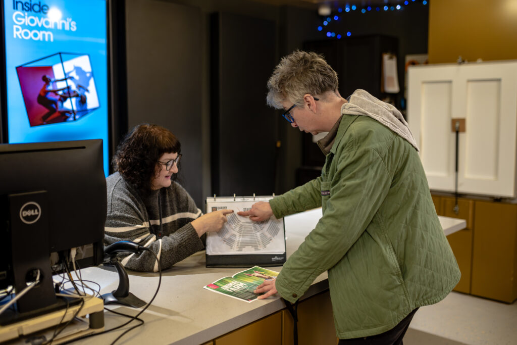 A visitor in a green jacket stands at a customer service desk. The customer assistant is holding and pointing at a seating plan. The visitor is also pointing at the seating plan.