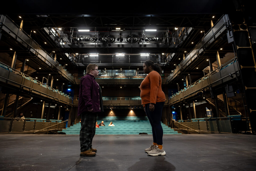 Two performers stand on stage with their eyes locked on each other. One person wears a sunflower lanyard. The auditorium is well lit and there is no audience.