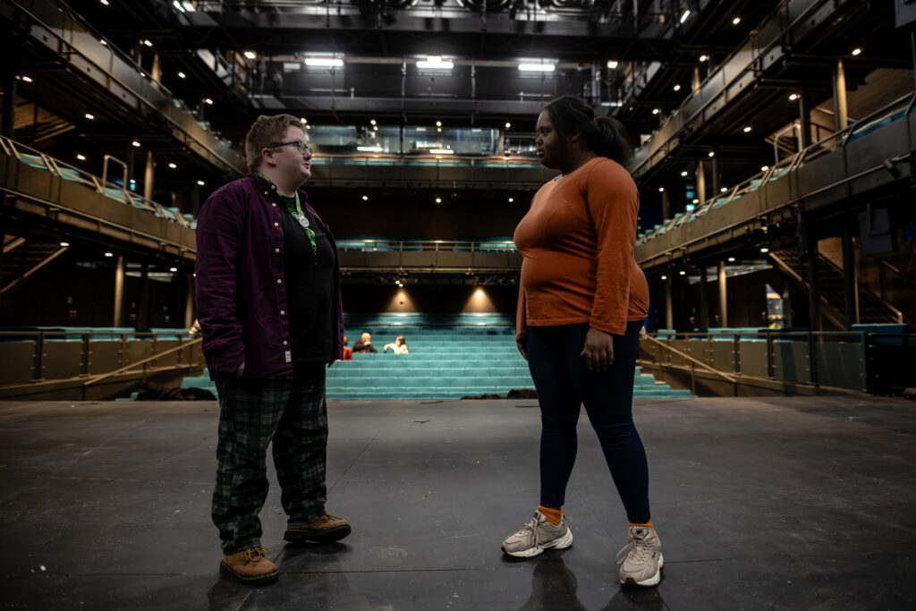 Two performers stand on stage with their eyes locked on each other. One person wears a sunflower lanyard. The auditorium is well lit and there is no audience.