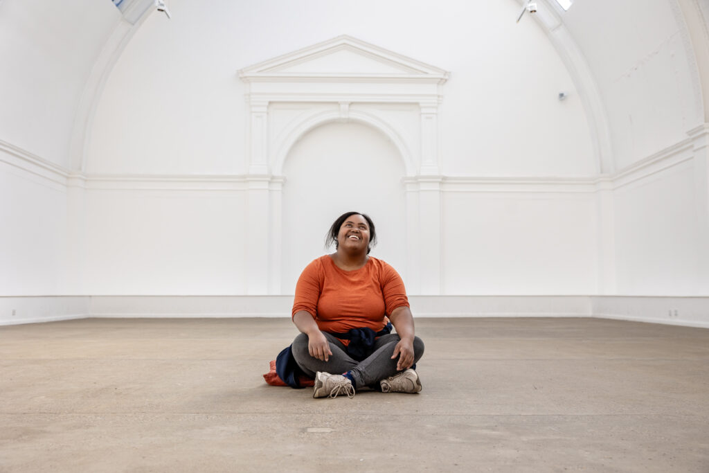 A person sits with their legs crossed on the floor on a clean, white gallery space with a curved ceiling. They look happy and optimistic.