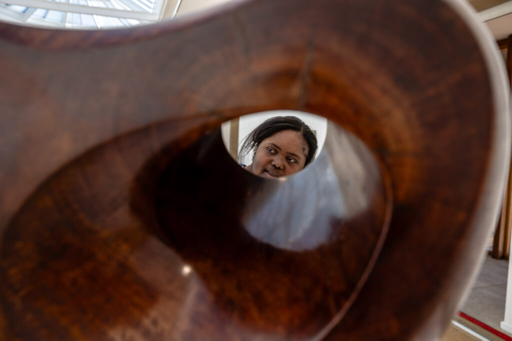 A person looks through a large, hollow wooden sculpture with smooth, curved surfaces.