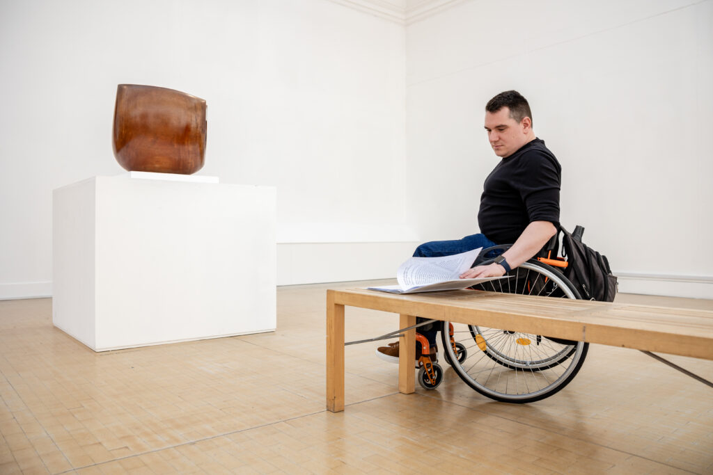 Person in a wheelchair in front of a wooden sculpture displayed on a white pedestal in a gallery setting. They are reading a document.