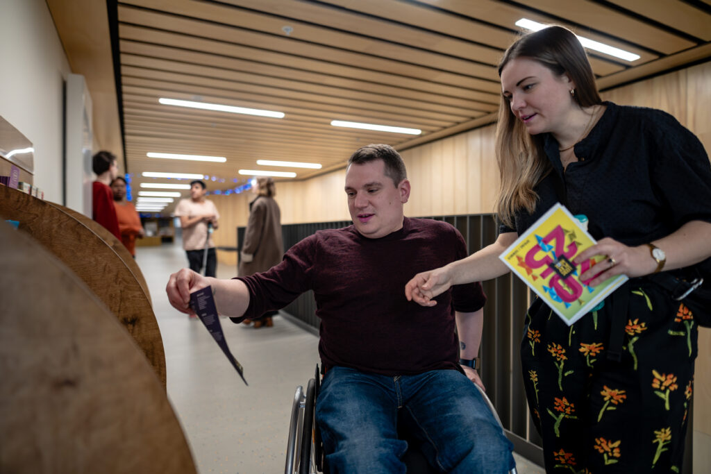 Two visitors look at a display of leaflets and brochures. One is a wheelchair. There are people stood talking in the background.
