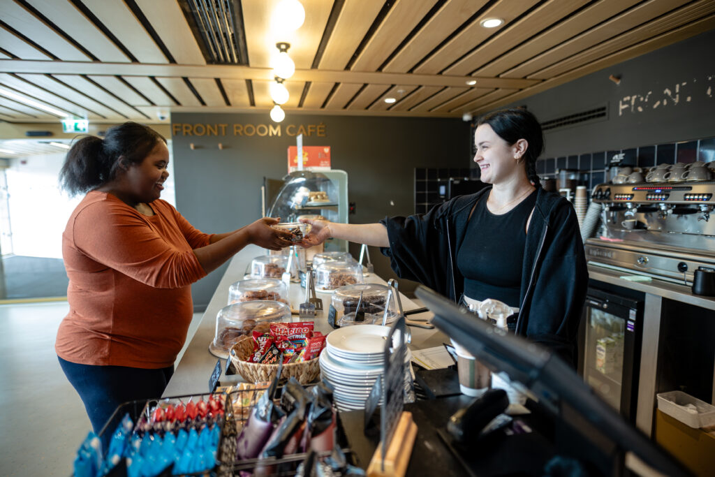 A staff member serves a visitor at a well-lit cafe counter that supports accessible communication.