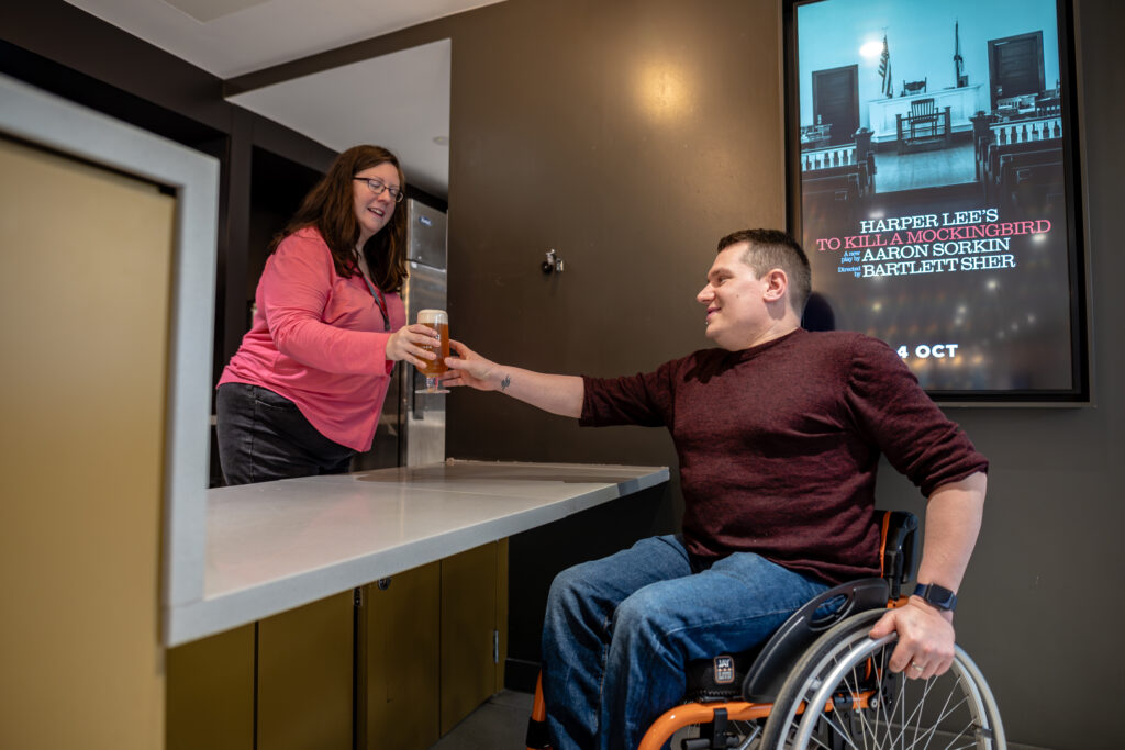 Wheelchair user receiving a drink across a lowered bar counter, with a poster for 'To Kill a Mockingbird' visible on the wall behind.
