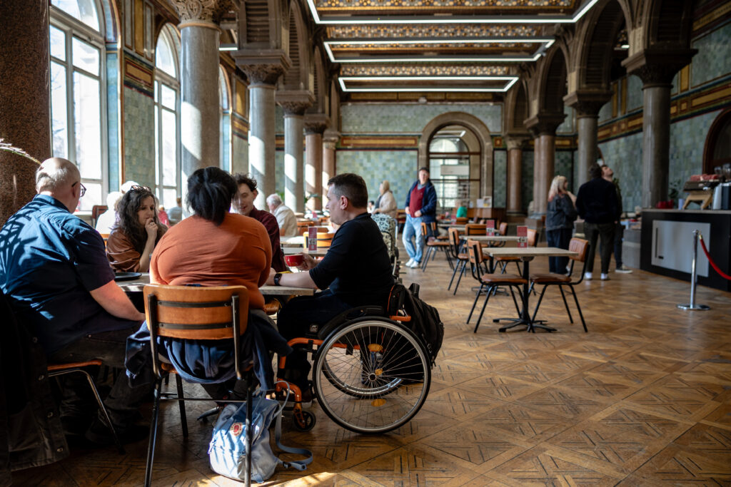 A group of people including a wheelchair user sit at a table in an ornate Victorian tearoom with a woodblock floor. 