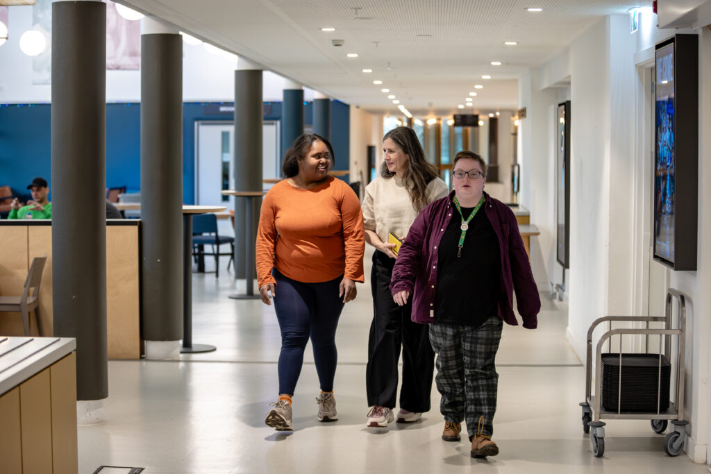 Three people are walking together down a wide, well-lit corridor with tables and chairs along the sides. A poster display is visible on the wall.
