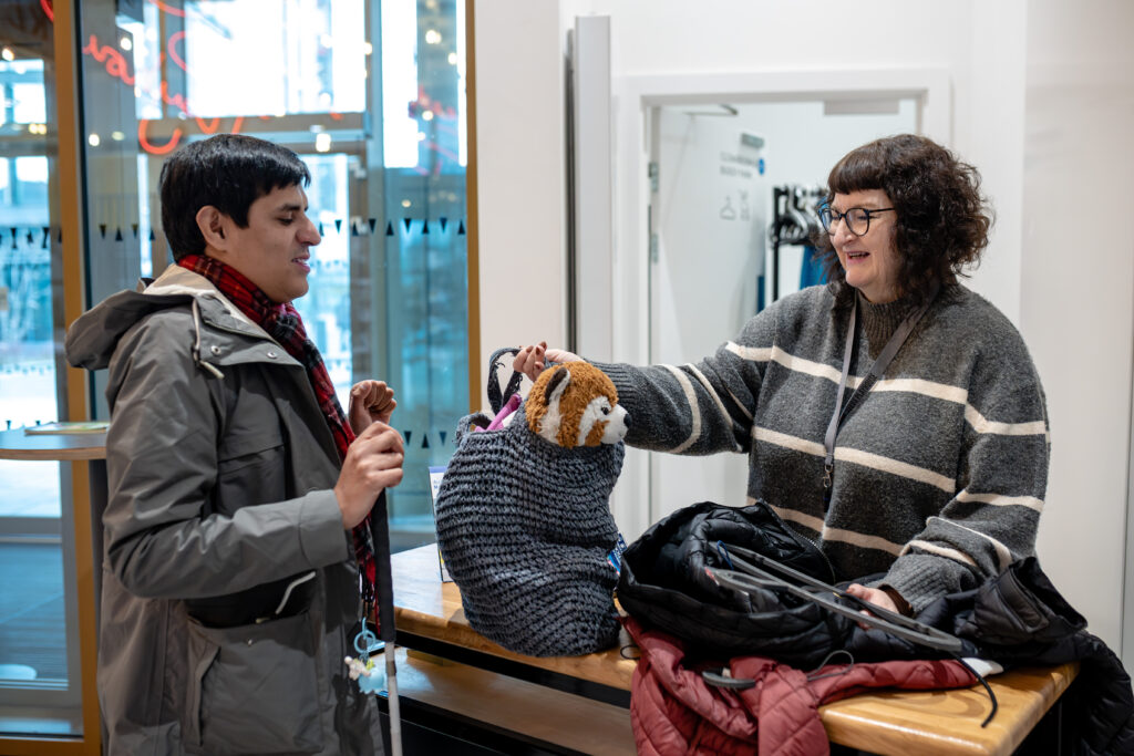 Two individuals are at a cloakroom counter. One person is holding a cane, whilst the member of staff is holding a large bag with a stuffed animal inside.