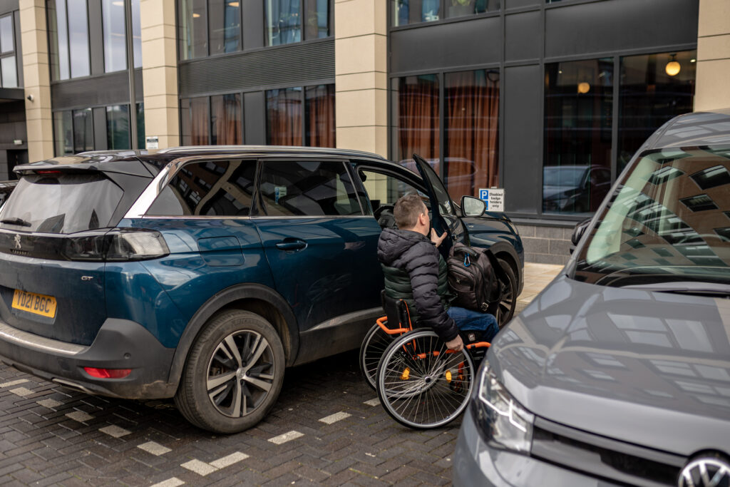 A wheelchair user gets out of a car in an accessible parking space in front of a venue.