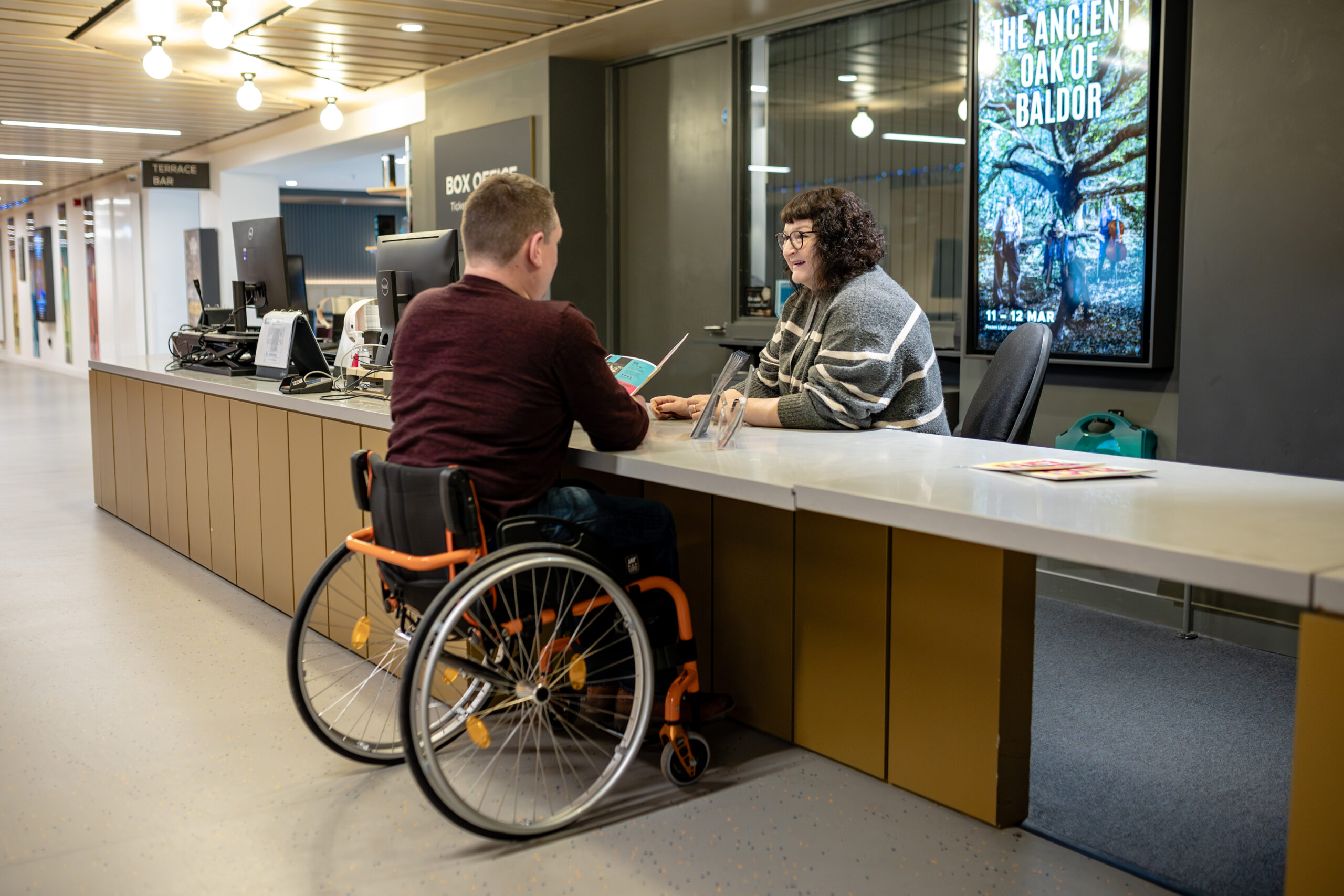 A wheelchair user is at a customer service counter, interacting with a staff member. There are computer monitors on the desk, and a poster on the wall that reads “The Ancient Oak of Baldor.”