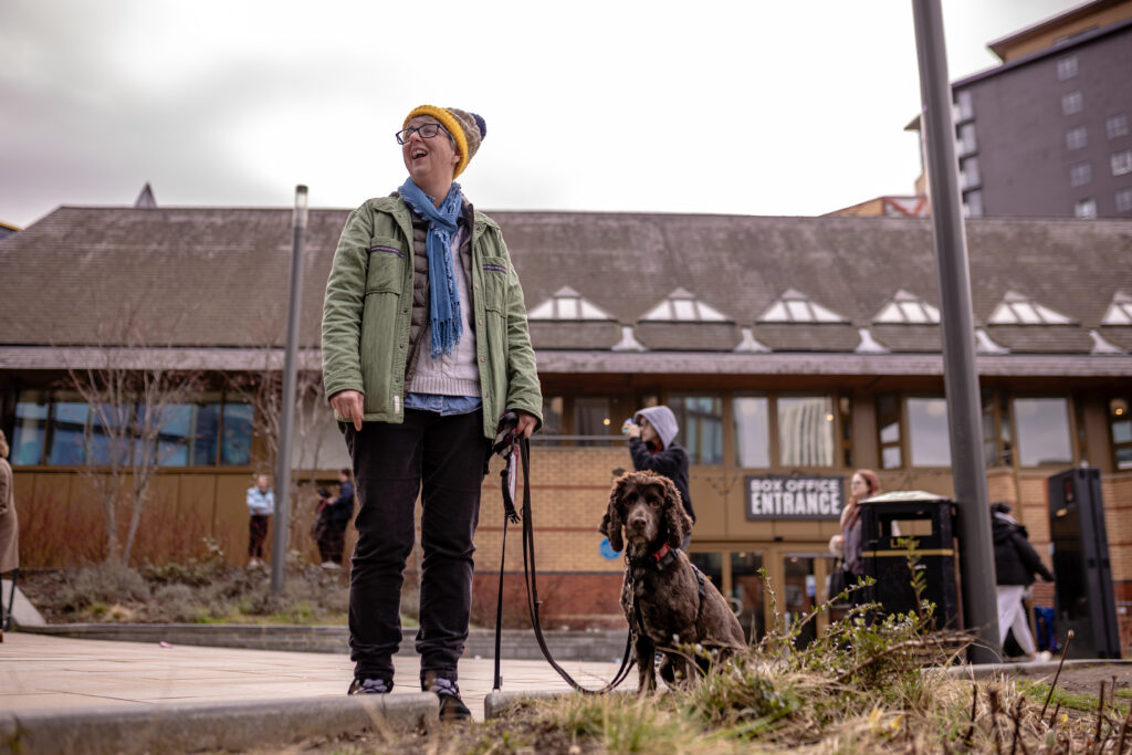 Assistance dog and owner in a flat spending area close to an arts venue. Streetlamps and a wastebin are nearby.