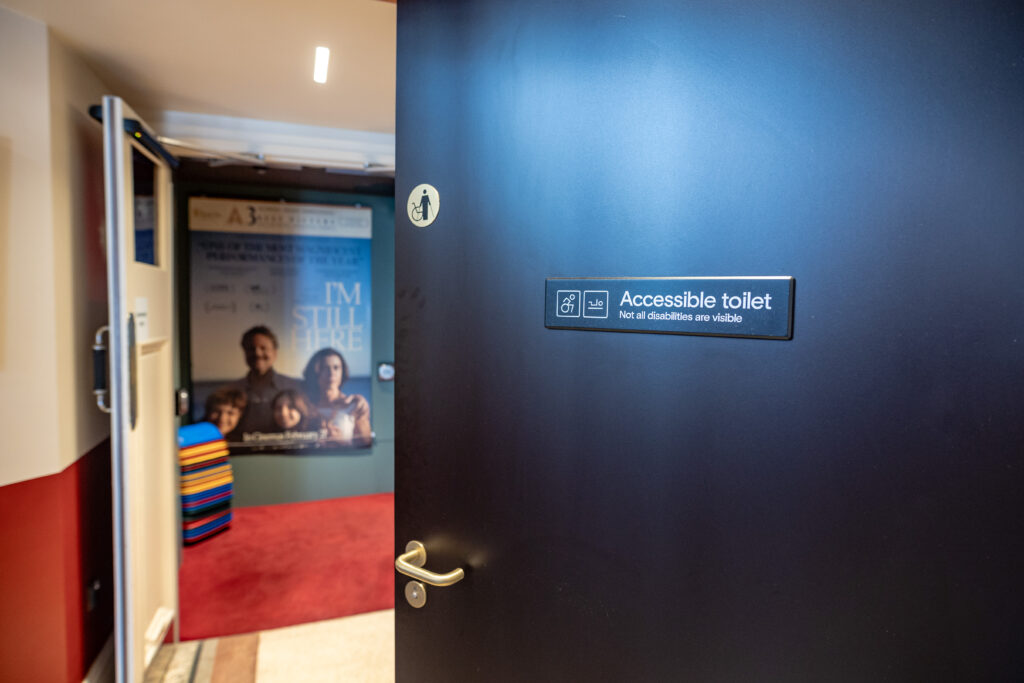 Open door with a sign reading 'Accessible toilet – Not all disabilities are visible. In the background there is a hallway with a red carpet and a poster on the wall