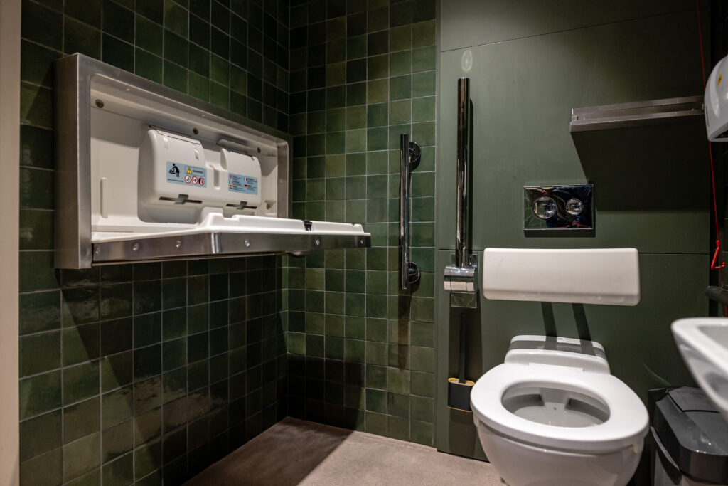 An open, wall-mounted baby changing unit inside an accessible toilet showing there is enough space for a wheelchair to turn.