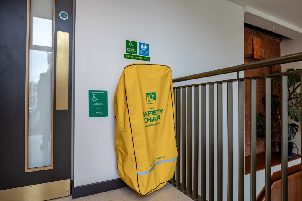 A bright yellow evacuation chair cover labeled “Safety Chair” is mounted on a wall near a staircase. Green emergency signage is visible above it.