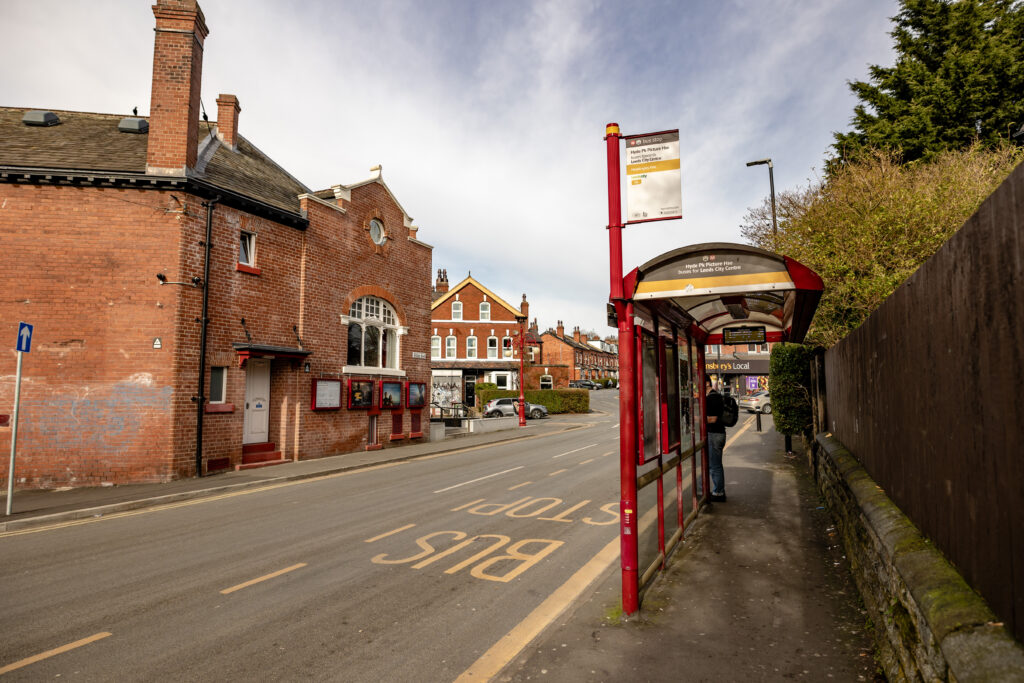 A red bus stop shelter on a quiet street lined with brick buildings and houses. The road has “BUS STOP” painted on it, and a timetable sign is visible on the shelter. Trees and a stone wall run along the right side of the pavement.