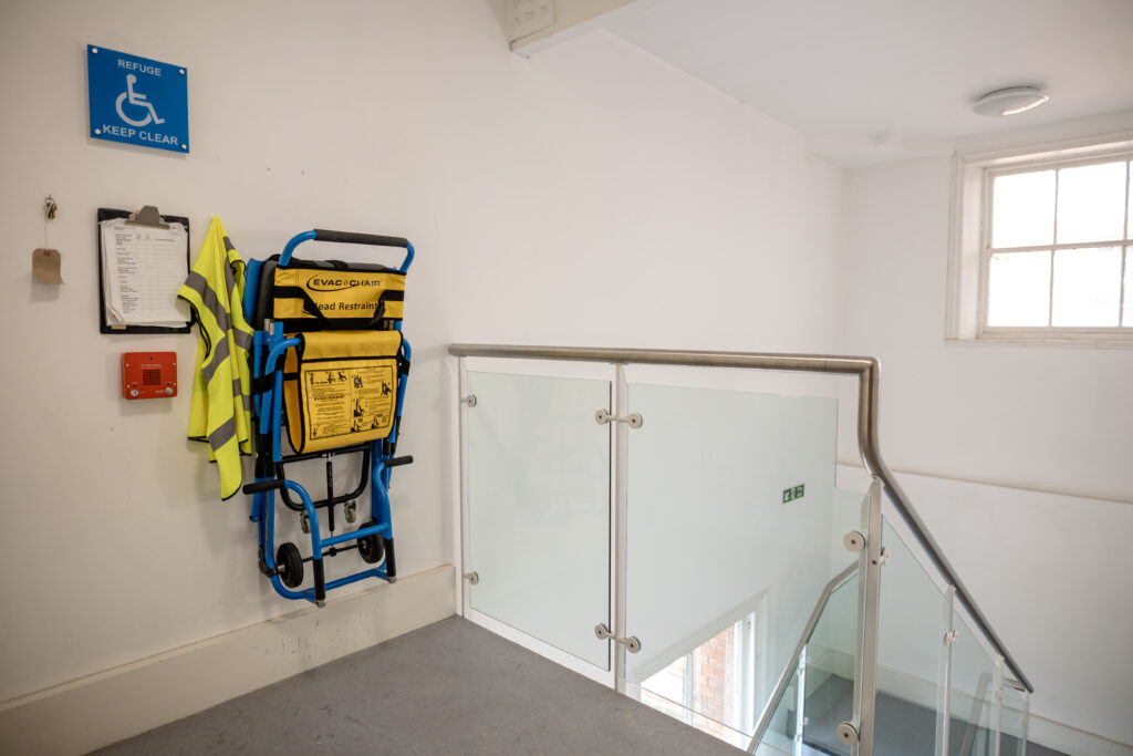 A refuge point on a stairwell with a blue ‘Refuge - Keep Clear’ sign, evac chair and accessible intercom button.