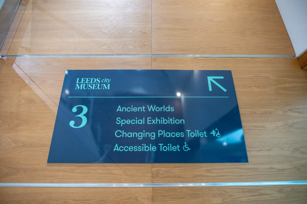 An accessible directional sign in a museum showing what facilities are on this level in clear, sans serif text with symbols.