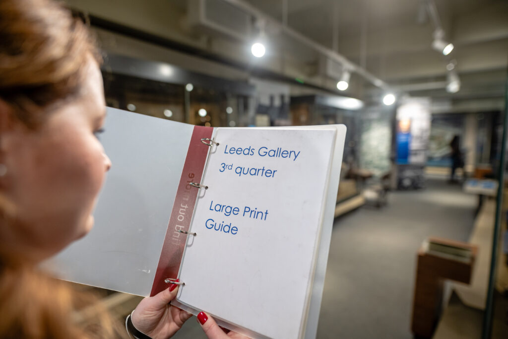Person holding a large print guide titled "Leeds Gallery 3rd quarter" in a museum setting.