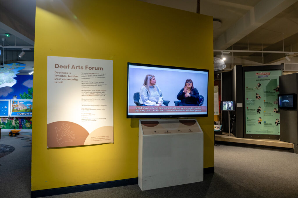 Exhibit display with a yellow wall featuring a Deaf Arts Forum information panel and a screen showing a video with captions about communicating with deaf people.