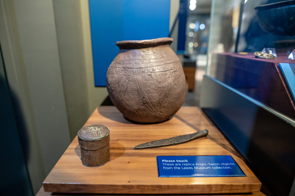 Display of replica Anglo-Saxon objects including a pot, a small container, and a knife on a wooden surface. There is a blue sign next to the objects which says ‘please touch’.