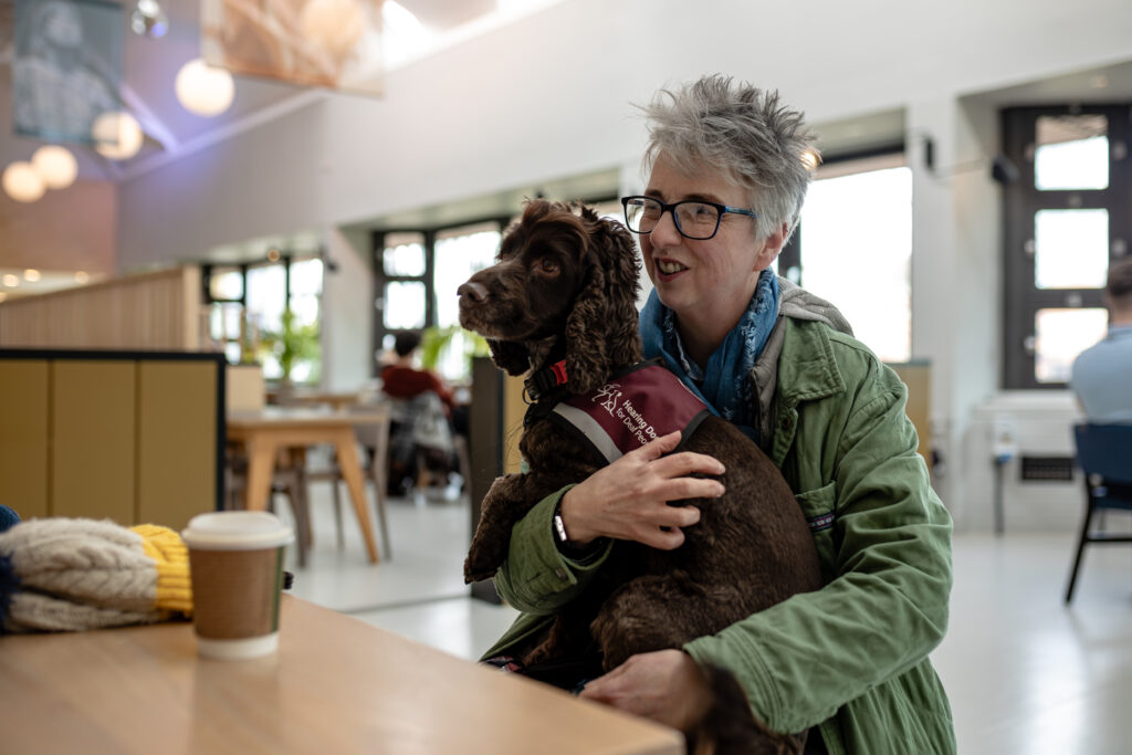 Person sitting indoors at a table with a coffee cup, holding a brown assistance dog wearing a coat that reads 'Assistance Dog'.