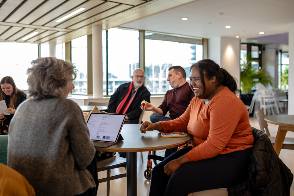 Group of people sitting around a table in a modern, bright indoor space with large windows, engaged in conversation and using a laptop.