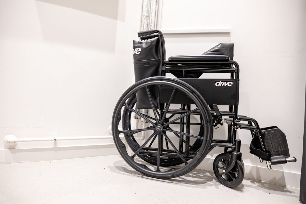 A black manual wheelchair is folded and placed against a white wall in a corridor.