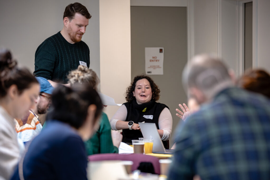 A group of people seated around a table engaged in a collaborative activity. Papers, notebooks, and drinks are on the table. One person is standing and holding a sheet of paper.