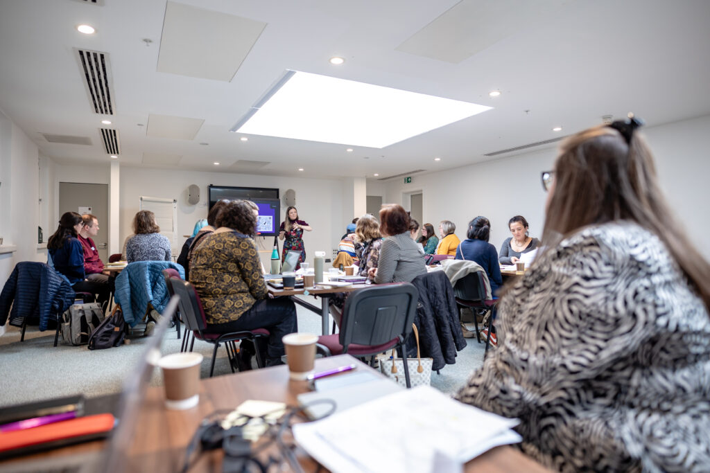 A person addresses a large group of people sat at round tables in a busy room.