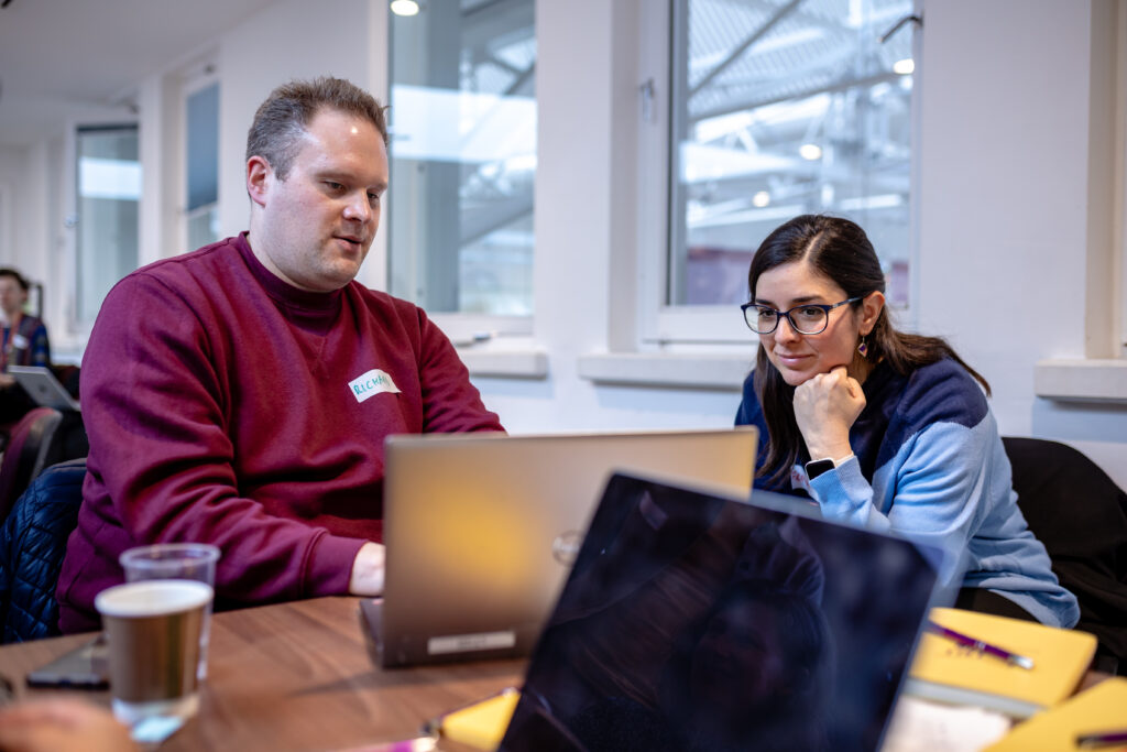 Two people sitting at a table with laptops in a bright indoor space, working together with notebooks and a drink nearby.